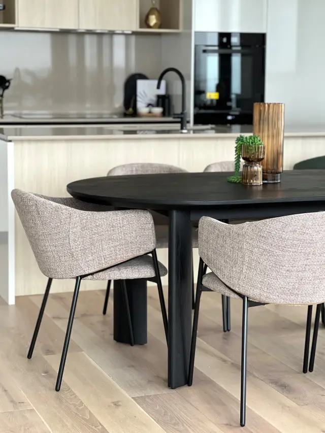 Dining area with black oval table, grey textured tub chairs and kitchen with black tapware beyond