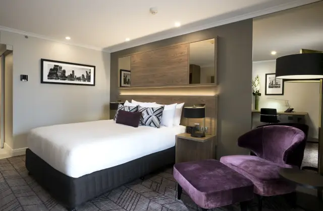 King guest room with backlit timber headboard, framed skyline artwork and aubergine velvet lounge chair
