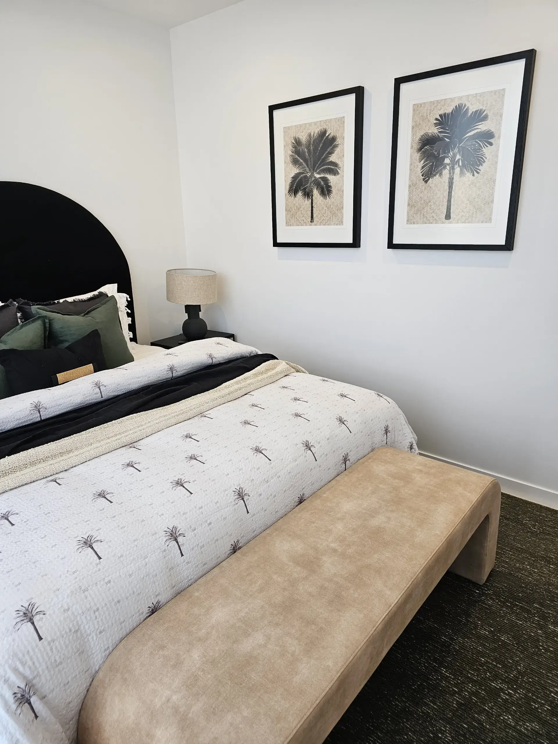 Bedroom in a Remi display suite with a black arched headboard, palm-print bedding, framed palm artwork and a tan bench at the foot of the bed
